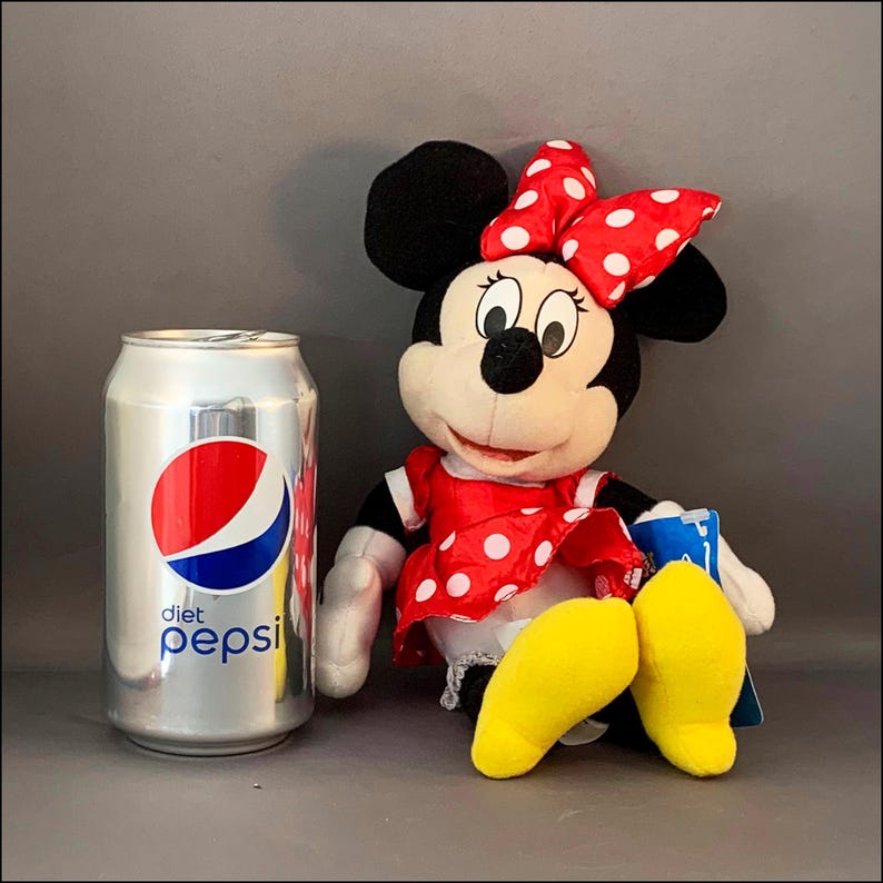 May include: A plush Minnie Mouse toy wearing a red dress with white polka dots and a red bow with white polka dots. The toy is sitting with its legs crossed and has yellow shoes. A can of Diet Pepsi is in the background.