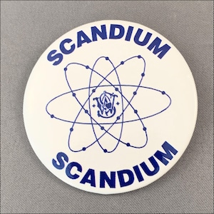 May include: A white button with the word "SCANDIUM" in blue, arched at the top and bottom. A blue atom symbol with a logo in the center is in the middle of the button.