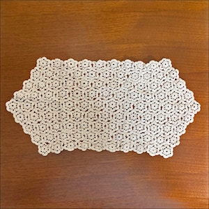 May include: Cream-colored, handmade lace doily with a floral pattern. The rectangular doily is made of delicate, open-weave lace, with a repeating pattern of small, six-petal flowers. The doily is placed on a wooden surface.