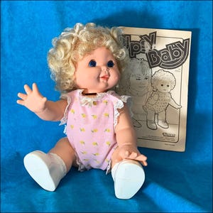 May include: A vintage 'Li'l  'Ups'y Baby' doll with blonde curly hair, blue eyes, and a pink and yellow floral print onesie. The doll is sitting on a blue surface with a vintage instruction booklet.