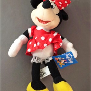 May include: A plush Minnie Mouse toy wearing a red polka dot dress with a white lace trim and a red bow. The toy has a white face with black eyes and a black nose. The toy is holding a blue tag that says "Disney".