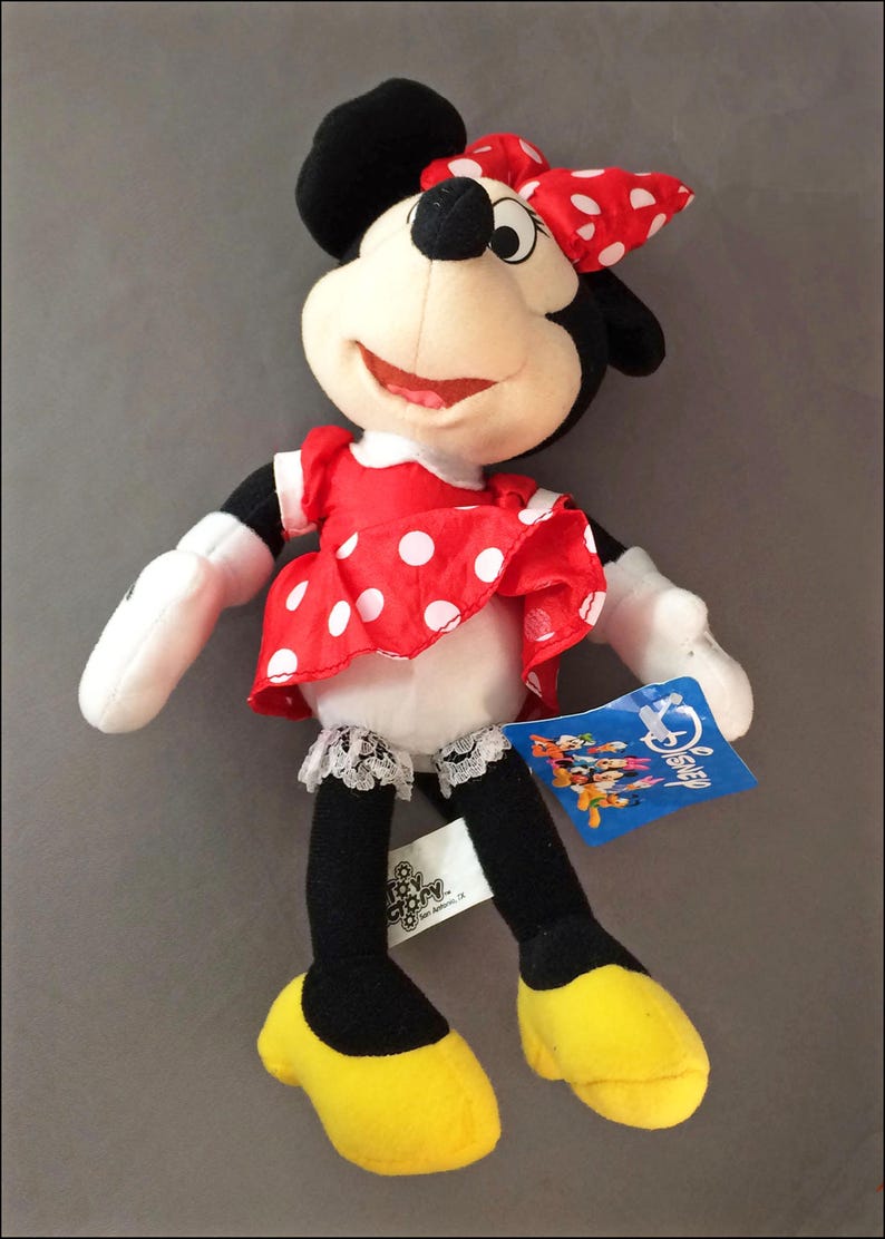 May include: A plush Minnie Mouse toy wearing a red polka dot dress with a white lace trim and a red bow. The toy has a white face with black eyes and a black nose. The toy is holding a blue tag that says "Disney".
