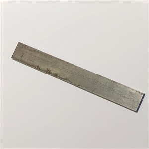 May include: A weathered, rectangular metal bar with a slightly rusted appearance. The bar is a dull silver color and lies diagonally across a white background. The metal shows signs of wear and age.