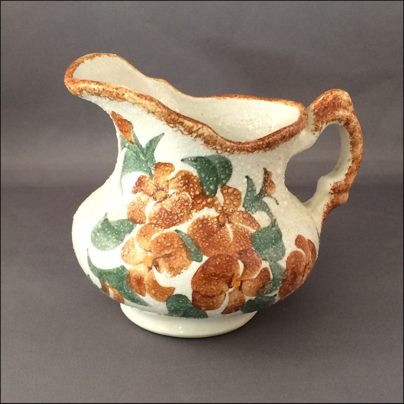May include: A white ceramic pitcher with a brown rim and handle. The pitcher is decorated with a floral pattern of orange and green flowers.