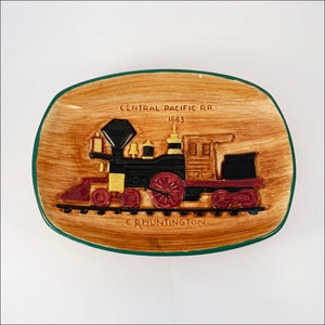 May include: A ceramic dish with a brown and green glaze depicting a vintage steam locomotive with the text "Central Pacific RR. 1863 C. Huntington".