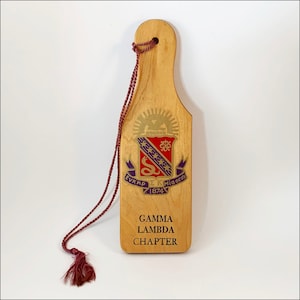 May include: A wooden paddle with a purple and gold crest and the text "ΕΥΚΑΡΠ ΣΚ Μια οδος 1874". The paddle also has the text "GAMMA LAMBDA CHAPTER" on the bottom.