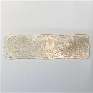 May include: A rectangular, off-white decorative object with an engraved design. The center features a circular emblem with two birds. The surrounding area is adorned with floral and leaf patterns. The object is likely a decorative element or a small, ornate plaque.