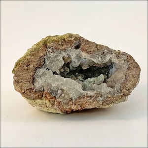 May include: A natural geode, cut open to reveal a sparkling interior. The outer shell is a rough, earthy brown with patches of green moss. The inside is lined with clear, crystalline formations, creating a cave-like appearance. The geode is displayed on a white surface.