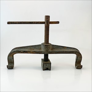 May include: An antique cast iron clamp with a threaded rod and handle. The clamp has a wide, curved base with two curved ends and a square base. The metal has a weathered, gray finish.