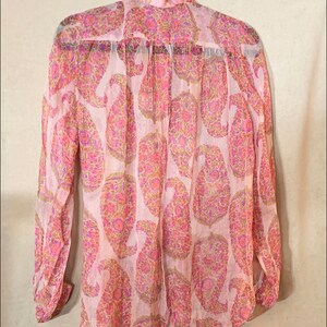 May include: A long-sleeved, pink, sheer blouse with a paisley print. The blouse has a button-up front and a gathered neckline.