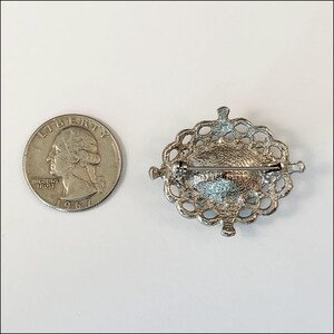 May include: Silver tone oval brooch with a filigree design and a pin on the back. The brooch is about the size of a quarter.