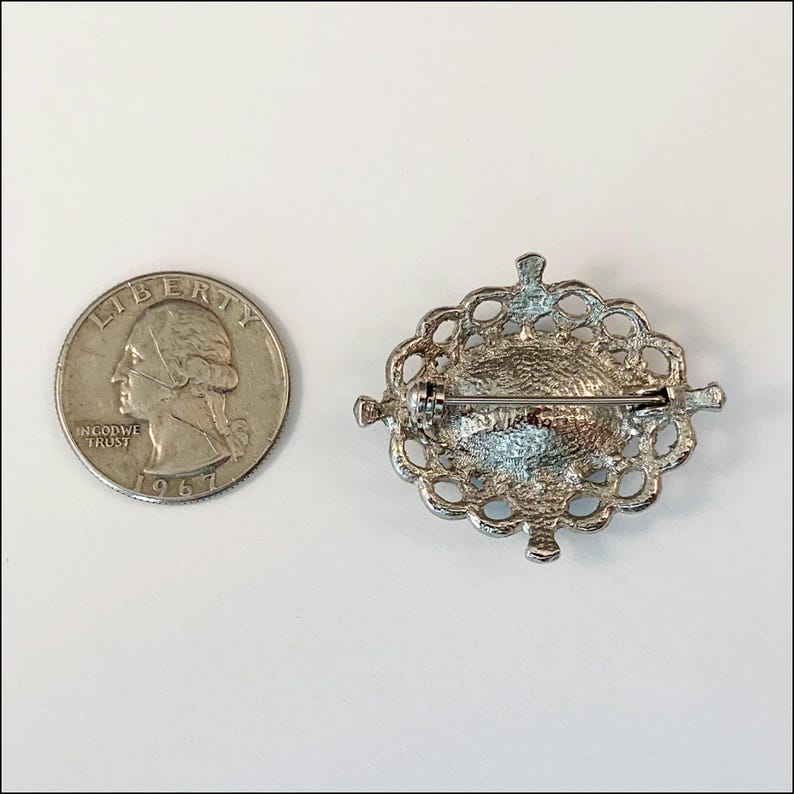 May include: Silver tone oval brooch with a filigree design and a pin on the back. The brooch is about the size of a quarter.