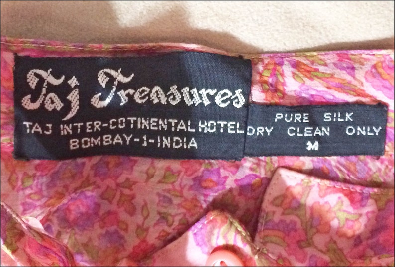 May include: A black label with white text that reads "Taj Treasures", "TAJ INTER-COTINENTAL HOTEL", "BOMBAY-1-INDIA", "PURE SILK", "DRY CLEAN ONLY", and "M".