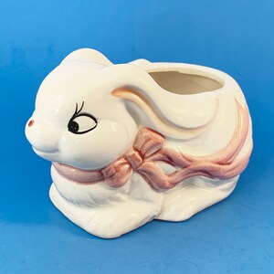 May include: White ceramic bunny planter with a pink ribbon. The bunny has black eyes and a small mouth.