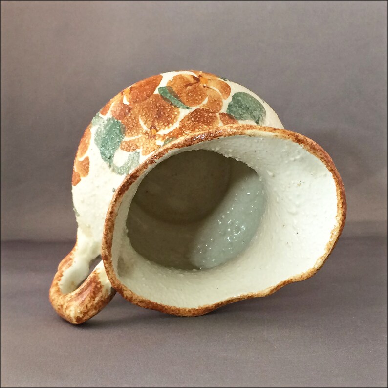 May include: A white ceramic pitcher with a brown and green floral design. The pitcher has a wide mouth and a handle.