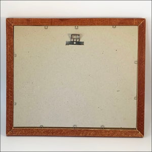 May include: A brown wooden picture frame with a light brown mat and a metal hanging hook on the back.