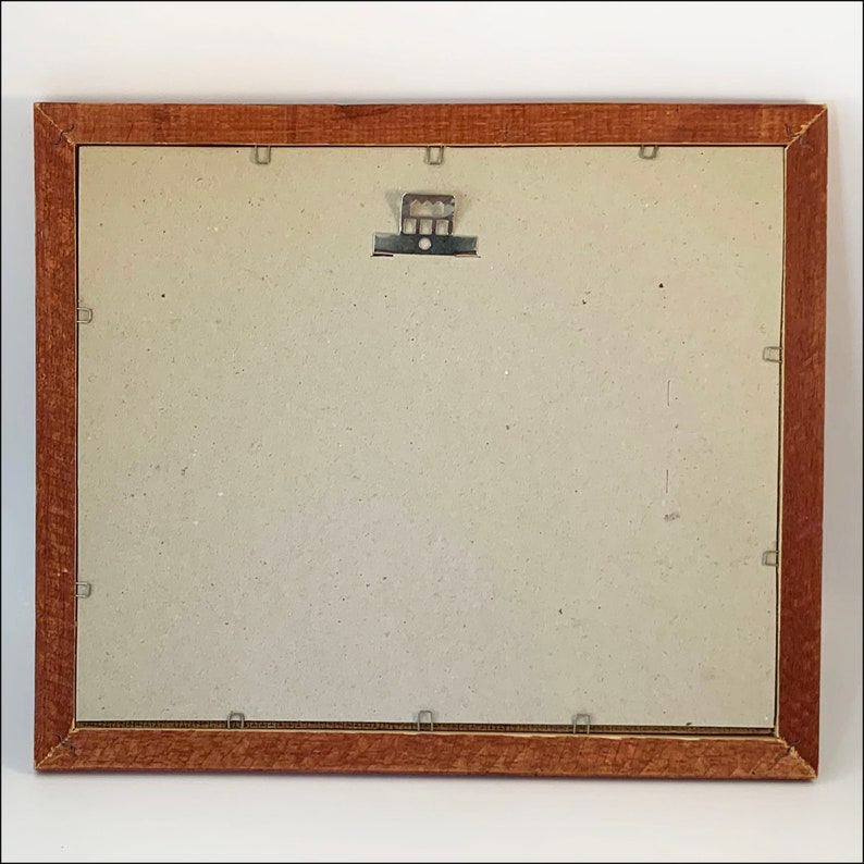 May include: A brown wooden picture frame with a light brown mat and a metal hanging hook on the back.