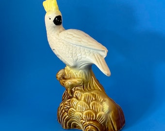 May include: A white ceramic cockatoo with a yellow crest perched on a brown ceramic tree stump.