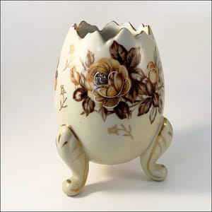 May include: A white ceramic egg-shaped vase with gold trim and a floral design featuring brown and yellow roses. The vase has three gold-painted feet.
