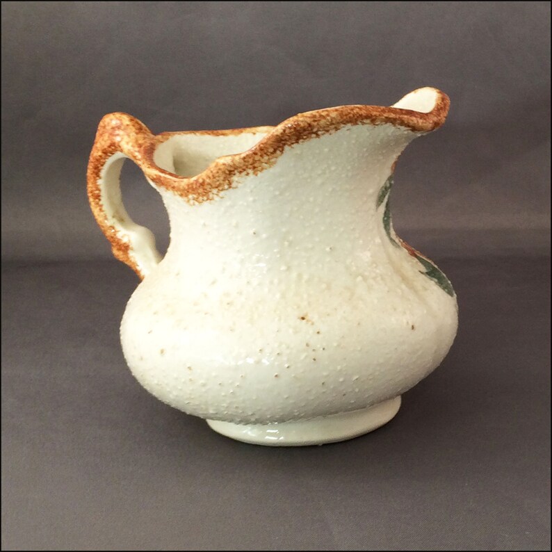 May include: A white ceramic pitcher with a brown rim and green floral design. The pitcher has a textured surface and a slightly flared lip.