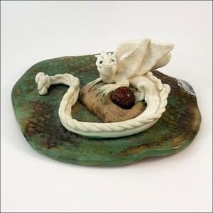 May include: A white ceramic dragon figurine with wings curled around a brown stone, sitting on a green and brown ceramic base.