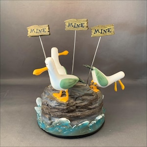 May include: A whimsical figurine featuring three seagulls perched on a rock formation with ocean wave details. Each seagull has a white body, green wings, and yellow beak and feet. Small wooden signs read "MINE".
