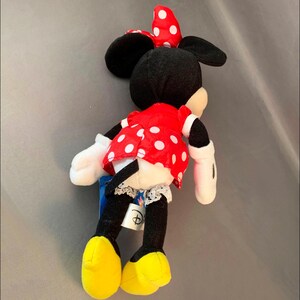 May include: A red and white polka dot dress with a black bow on top of the head. The plush toy is a Minnie Mouse character with black ears and yellow shoes.