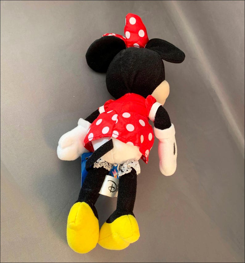May include: A red and white polka dot dress with a black bow on top of the head. The plush toy is a Minnie Mouse character with black ears and yellow shoes.