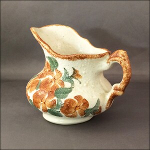 May include: A white ceramic pitcher with a brown and green floral design. The pitcher has a scalloped rim and a handle.