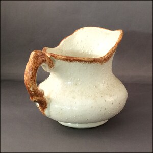 May include: A white ceramic pitcher with a brown handle and a textured surface. The pitcher has a wide, rounded base and a flared lip.