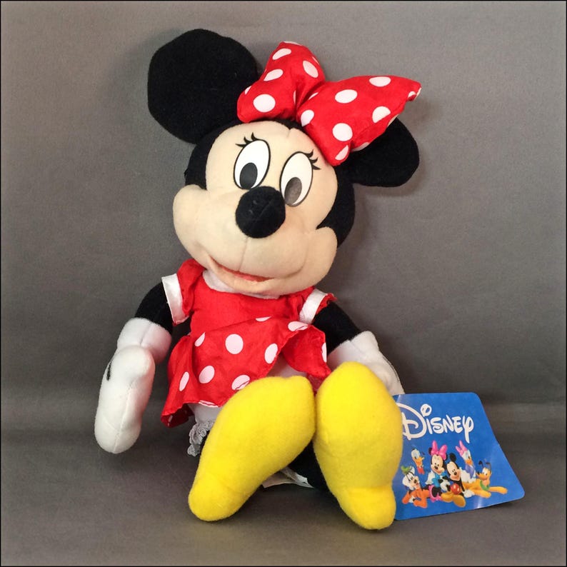 May include: A plush Minnie Mouse toy wearing a red dress with white polka dots and a red bow with white polka dots. The toy has yellow feet and is sitting with its legs crossed. The toy has a Disney tag attached to it.