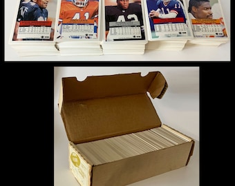 May include: A brown cardboard box with a white label that says "Pro Set Football Cards" is open and filled with stacks of white trading cards. The cards feature photos of football players, including Steve Broussard, Greg Lewis, Ronnie Lott, Phil Simms, and Gary Anderson.