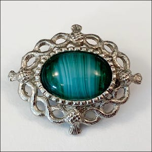 May include: A silver brooch with a green and white agate stone set in a Celtic knot design. The brooch is decorated with four thistle motifs.