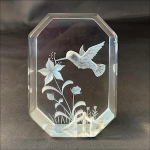 May include: A clear, octagonal glass paperweight featuring an etched design of a hummingbird in flight approaching a large five-petaled flower. The base of the scene includes smaller buds, grass-like stems, and a small fence-like structure.