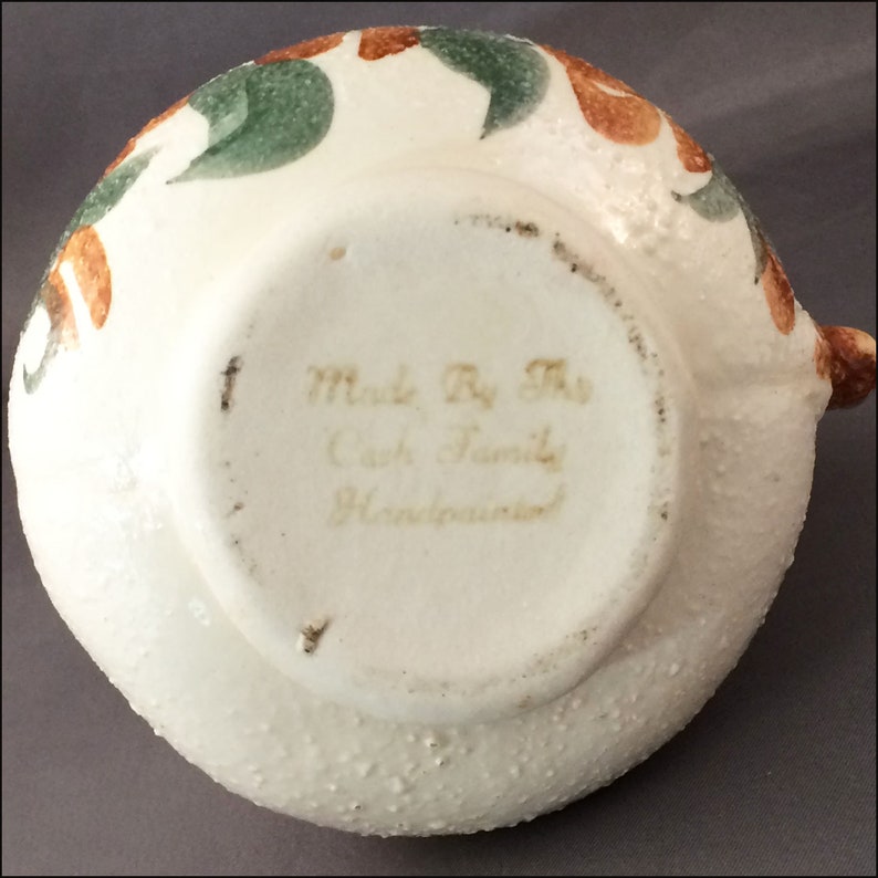 May include: White ceramic pitcher with a brown and green floral design. The bottom of the pitcher reads "Made By The Cash Family Handcrafted".