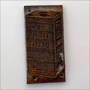 May include: A vintage metal printing block with the text "OF LEAD ARSENATE DOW" and a depiction of a can. The block has a rough, textured surface and is brown in color.