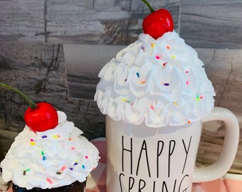 May include: A white ceramic mug with the text HAPPY SPRING in black lettering, topped with faux white whipped cream, colorful sprinkles, and a red cherry. Beside it sits a small white marshmallow-shaped mug with a face and a cherry.