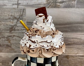 May include: A black and white checkered mug filled with a faux hot cocoa beverage. The drink is topped with swirls of brown and white whipped cream, miniature white marshmallows, a small chocolate bar, and a gold straw.
