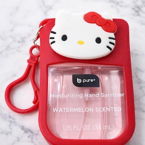 May include: Red hand sanitizer holder with a white Hello Kitty face and red bow. The holder contains a clear bottle of watermelon-scented hand sanitizer. The bottle is 1.15 fl oz (34 ml) and has a red clip.