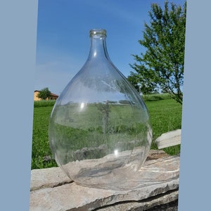 May include: A large, clear glass demijohn bottle with a wide, rounded body and a narrow neck. The bottle sits on a stack of gray stone slabs, with a bright blue sky and green grass in the background.