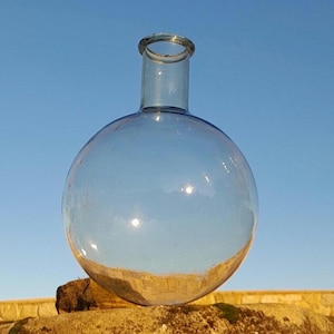 May include: A clear glass round bottle with a narrow neck sits on a brown rock with green moss.