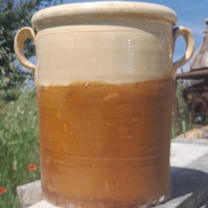 May include: A large, cylindrical ceramic pot with two curved handles. The pot has a two-tone color scheme, with a light beige top and a darker brown lower section. The pot is outdoors, set against a backdrop of a blue sky and greenery.