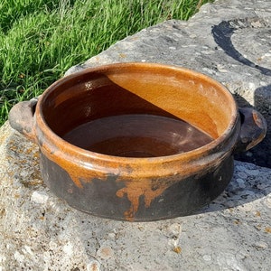 Puede incluir: Una olla de barro marrón con dos asas se asienta sobre una gran roca gris texturizada. La olla está llena de agua y tiene un esmalte marrón oscuro en el fondo.