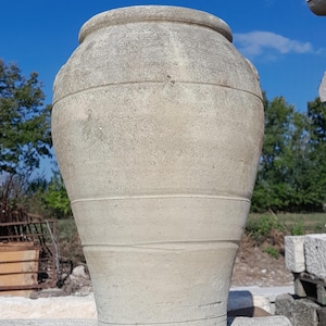 Könnte beinhalten: Ein großer, verwitterter, beigefarbener Keramiktopf mit breiter Öffnung und rundem Körper. Der Topf hat horizontale Bänder und steht auf einer Steinoberfläche vor blauem Himmel.