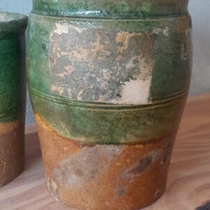 May include: A pair of antique ceramic pots with a distressed finish. The pots are cylindrical with a wide rim and are painted in shades of green and brown. The pots are likely for decorative use.
