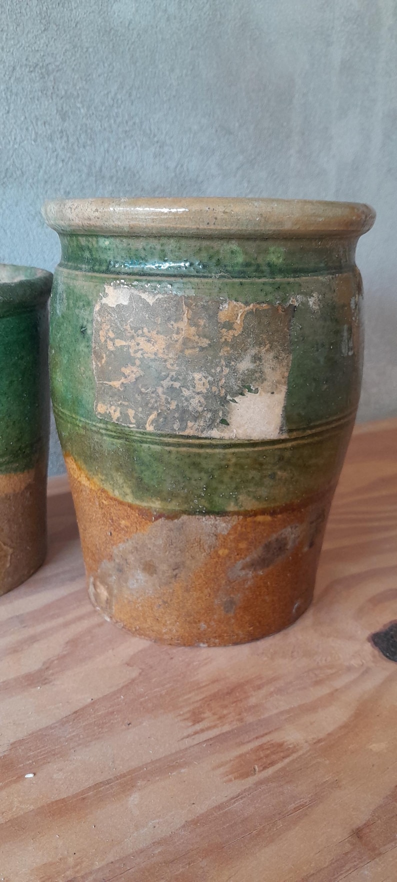 May include: A pair of antique ceramic pots with a distressed finish. The pots are cylindrical with a wide rim and are painted in shades of green and brown. The pots are likely for decorative use.
