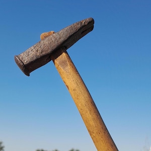 May include: A vintage hammer with a weathered, rusty metal head and a long, wooden handle. The hammer is held against a clear blue sky, with a backdrop of green trees and dry grass.