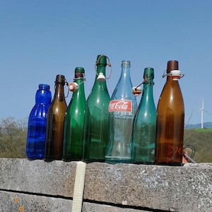 Op de afbeelding: Een verzameling van zeven glazen flessen in verschillende kleuren en vormen. De flessen hebben kurken en sommige hebben draadsluitingen. De flessen zijn in een rij op een betonnen oppervlak gerangschikt.