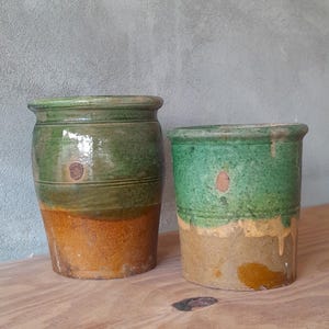 May include: Two antique ceramic pots with a rustic appearance. The pots are cylindrical, with a green upper section and a tan lower section. The pots are displayed on a wooden surface against a grey wall.