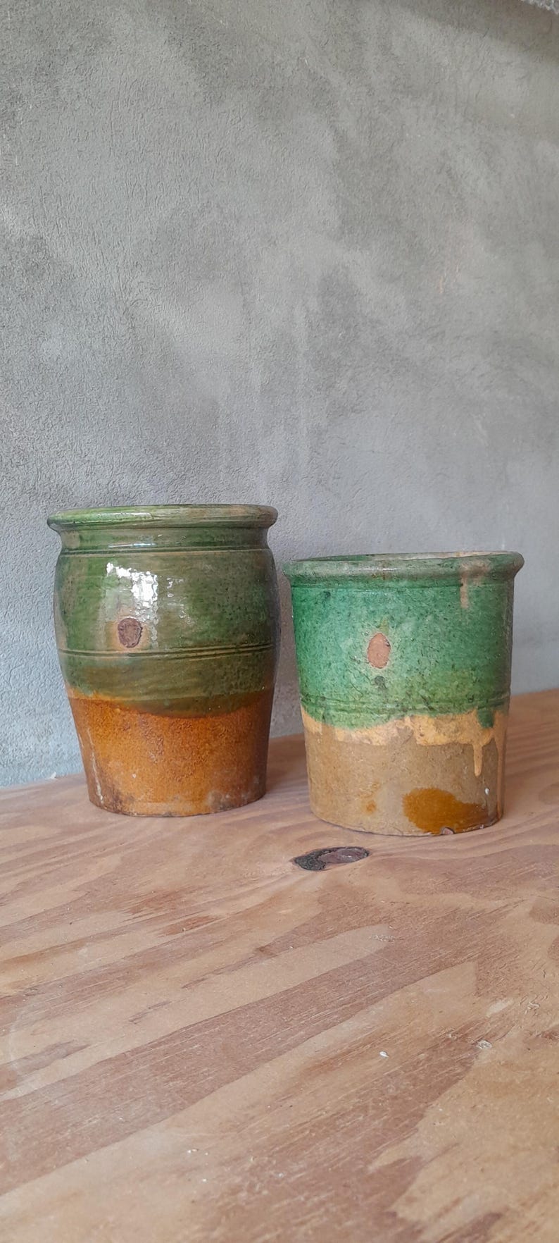 May include: Two antique ceramic pots with a rustic appearance. The pots are cylindrical, with a green upper section and a tan lower section. The pots are displayed on a wooden surface against a grey wall.